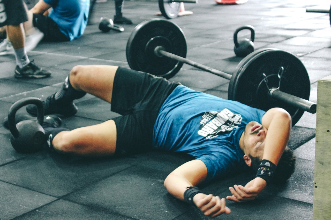Atleta com a mão sobre o rosto e deitado no chão ao lado de uma barra reto com anilhas grandes. O atleta parece estar exausto.
