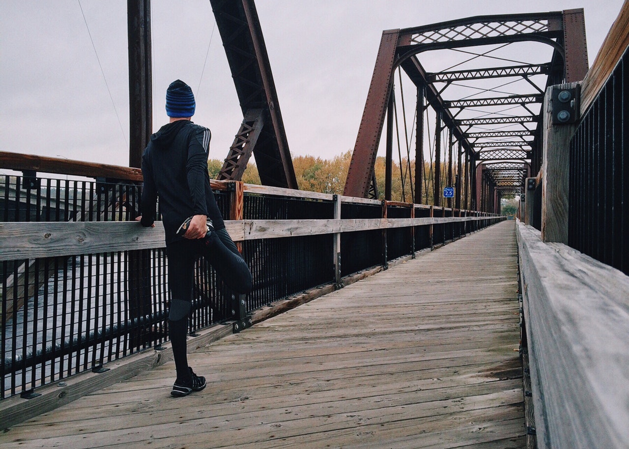 Imagem de um atleta em uma ponte se alongando para começar uma caminhada, parece estar frio no ambiente