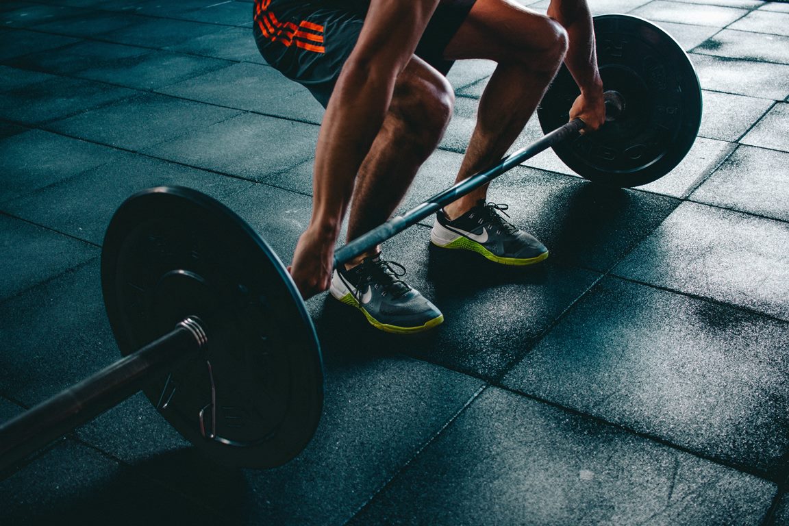 Imagem de um atleta com uma barra com pesos em suas extremidades. O atleta está segurando a barra e com os joelhos flexionados.
